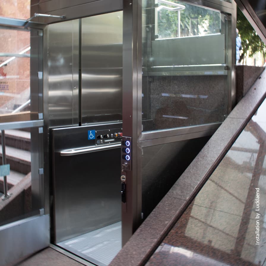 V-1504 Lux vertical platform lift with brushed stainless steel enclosure at commercial building entrance